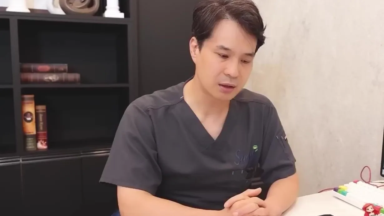 회색 수술복을 입은 의사가 책장 앞 책상에 앉아 아래를 보며 말하는 모습