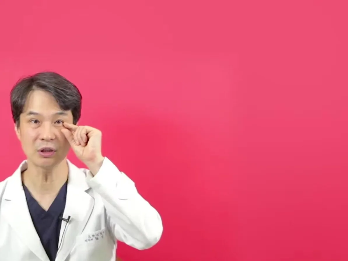 흰 가운을 입은 의사가 손가락으로 눈꼬리 쪽을 가리키며 설명하는 모습